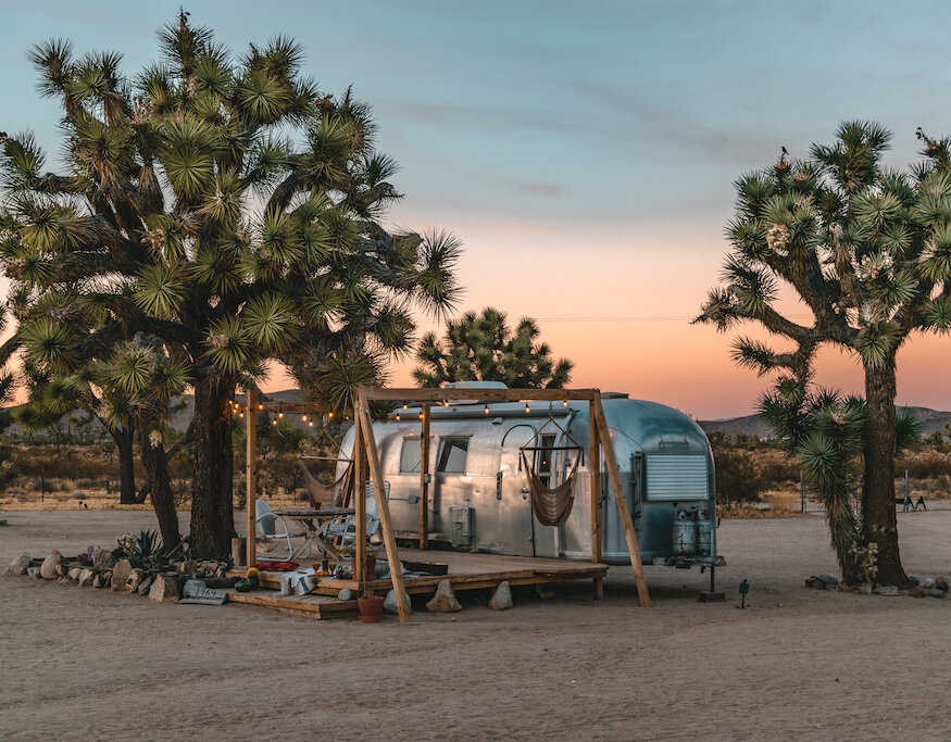 An Airstream under Joshua trees at dusk, with a deck and string lights